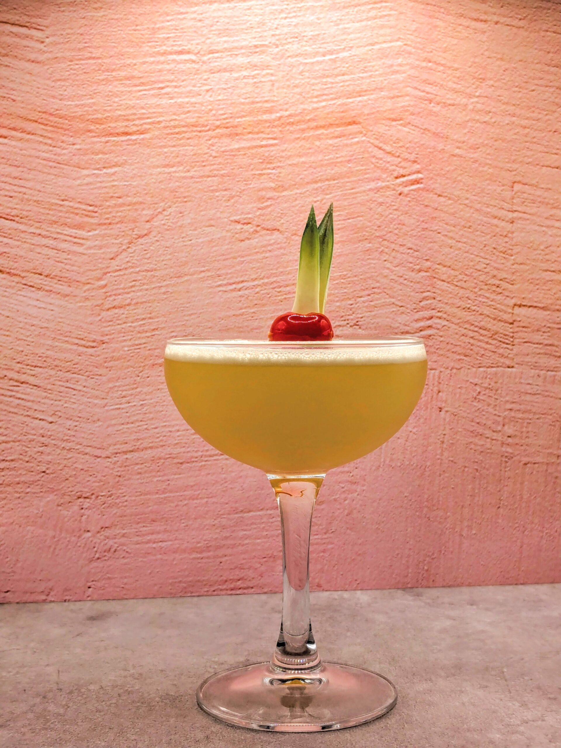 A pale golden cocktail in a couple glass, garnished with a cherry against a pink backdrop